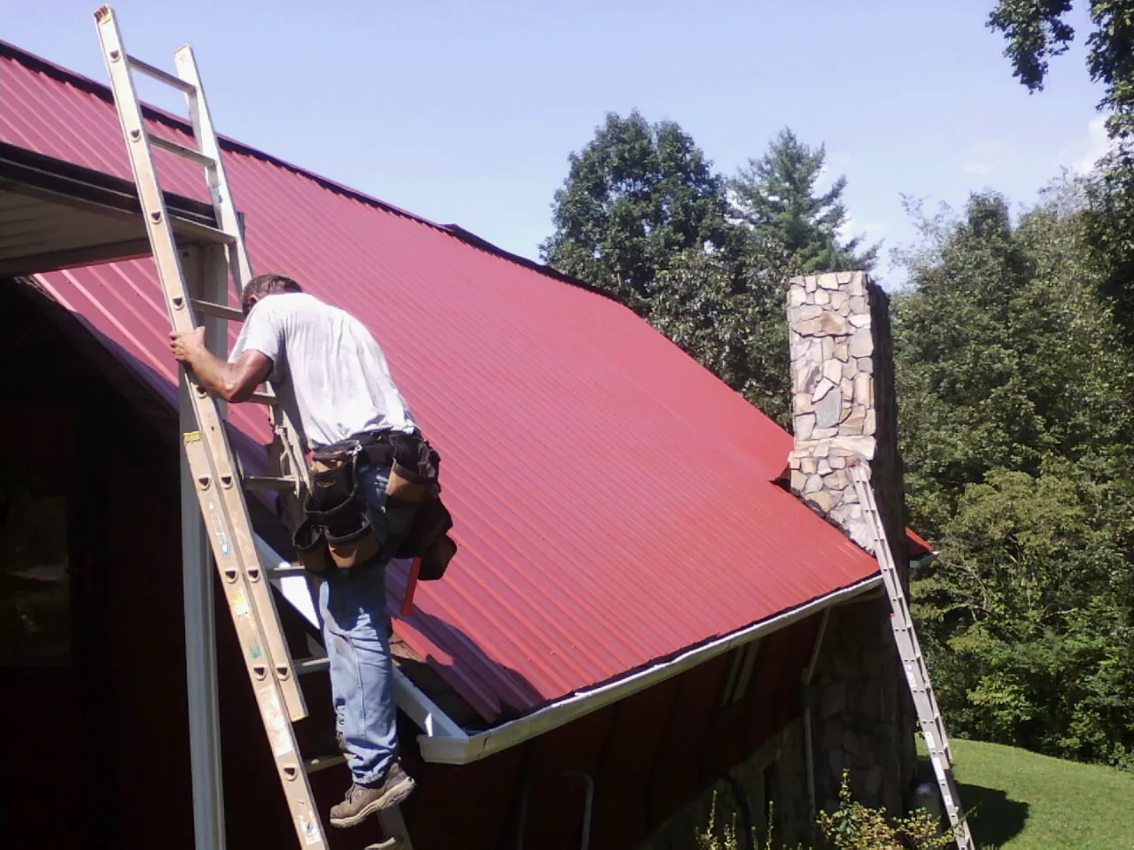 Roofer on a ladder beside a red metal roof and stone chimney — Soffit & Fascia Repair by Roofing Brothers in Monterey